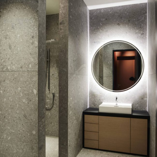 Bad mit Terrazzo-Wänden, beleuchtetem Rundspiegel und Holzwaschtisch im Business Apartment
