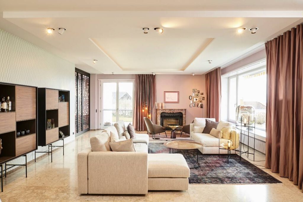 Großer Wohnraum mit hellem Sofa, Perserteppich, Kamin und roséfarbenen Vorhängen in der Villa A