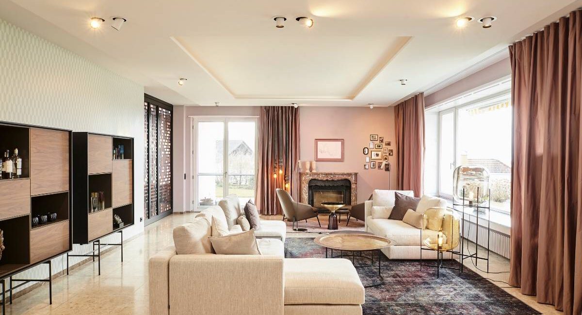 Großer Wohnraum mit hellem Sofa, Perserteppich, Kamin und roséfarbenen Vorhängen in der Villa A