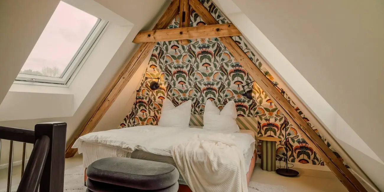 Schlafzimmer unter Dachbalken mit gemusterter Tapete und Samthocker in der Villa Haag