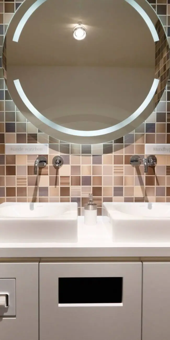 Doppelwaschtisch mit beleuchtetem Rundspiegel vor braunem Mosaik in der Praxis Winner