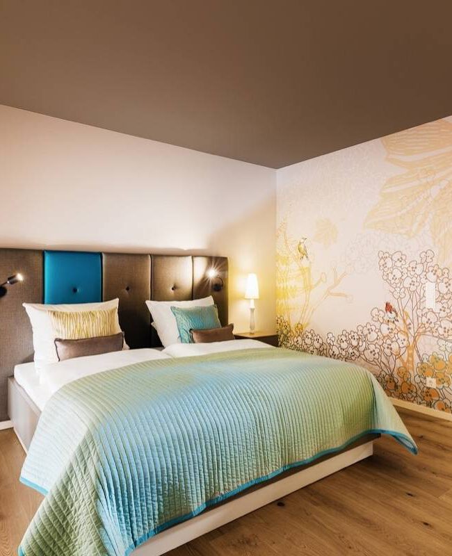 Doppelbett mit petrolfarbenem Kopfteil und Wandmalerei im Hotelzimmer Taufstein