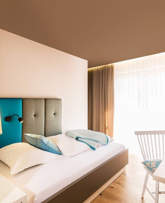 Doppelbett mit türkisen Kissen und Schreibtisch am Fenster im Hotelzimmer Taufstein