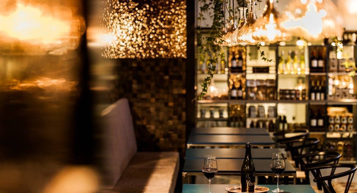 Gedeckter Tisch mit Rotweingläsern vor warm beleuchtetem Weinregal in Brunos Bistro