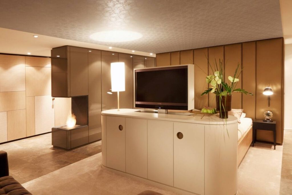 Freistehendes Sideboard mit Fernseher vor goldfarbener Wandverkleidung und Kaminfeuer in der Suite A