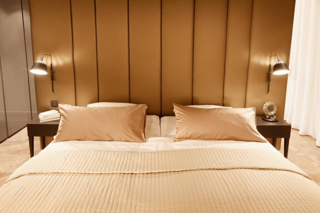 Doppelbett vor gepolsterter Wandverkleidung in Goldtönen mit zwei Wandleuchten in der Suite A