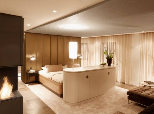 Schlafbereich mit Kaminblock, Polsterwand in Goldtönen und Vorhängen in der Suite A