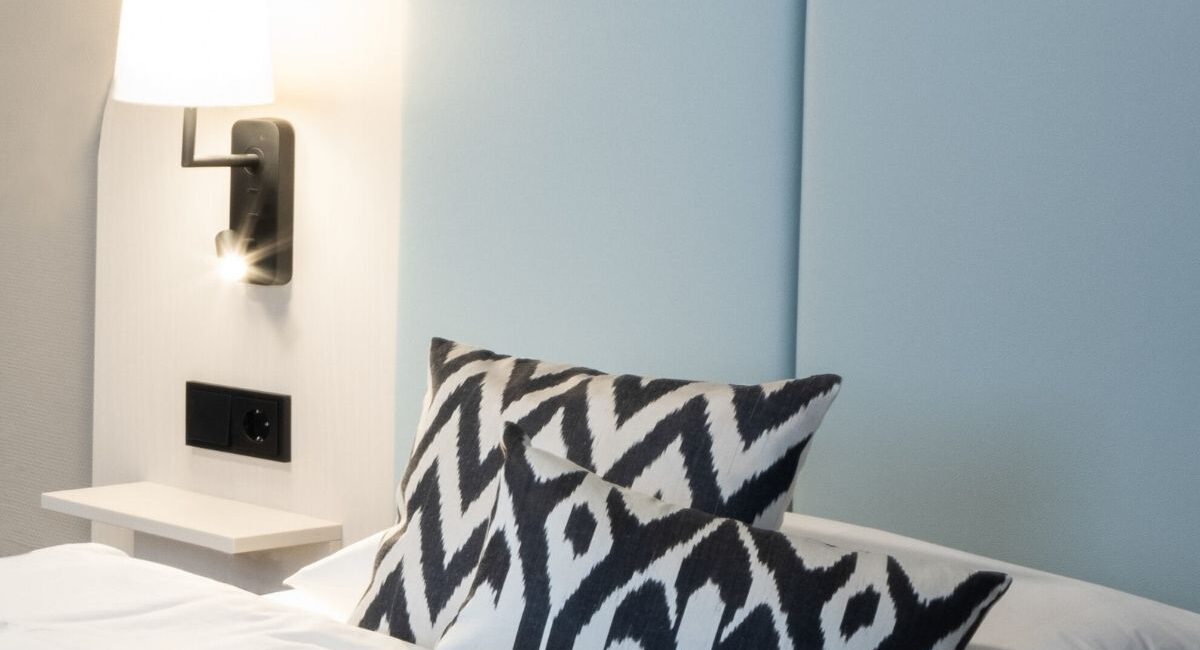 Bett mit hellblauem Kopfteil und Ikat-Kissen unter Wandleuchte, Haus der Standesbeamten Haus 3