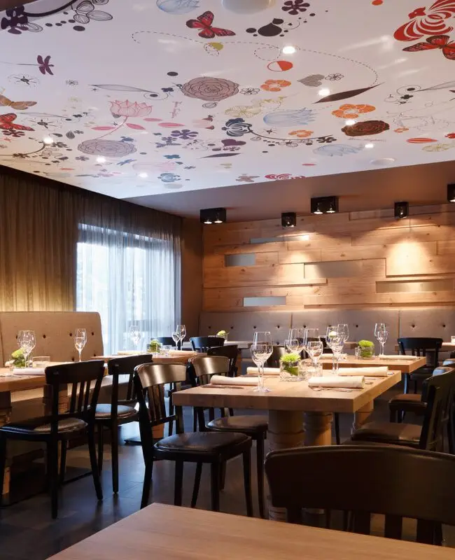 Gastraum mit bunt bedruckter Decke, Holzwand und dunklen Stühlen im Hotel Taufstein Restaurant I