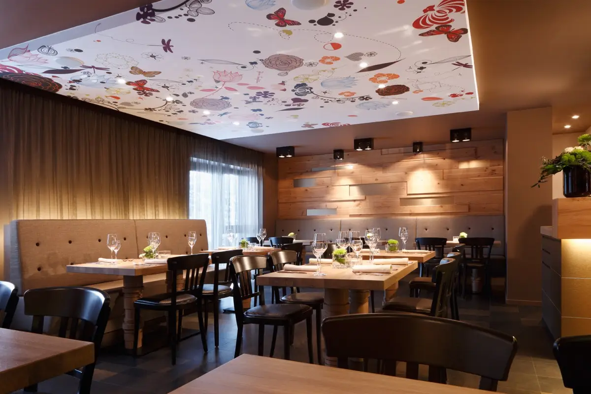 Gastraum mit bunt bedruckter Decke, Holzwand und dunklen Stühlen im Hotel Taufstein Restaurant I